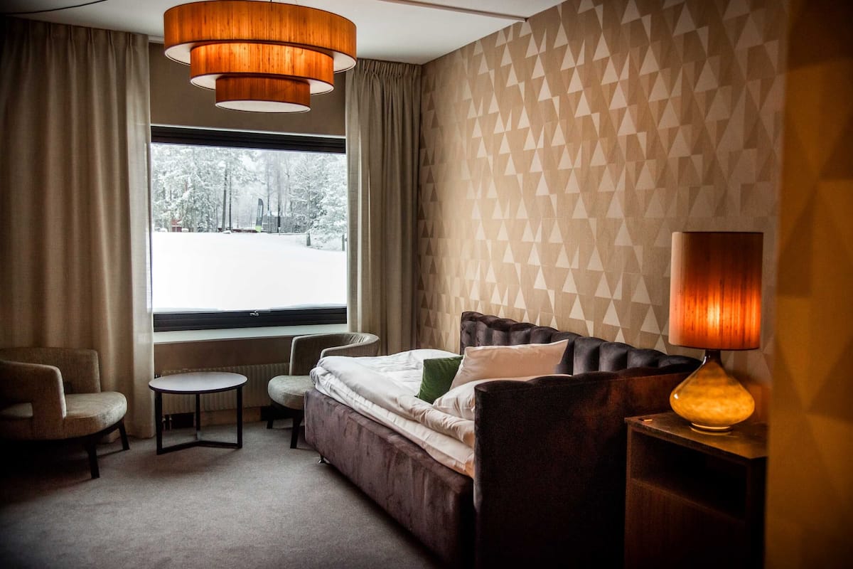 Une chambre d’hôtel avec un lit, deux chaises, une petite table, une lampe et une fenêtre donnant sur des arbres.