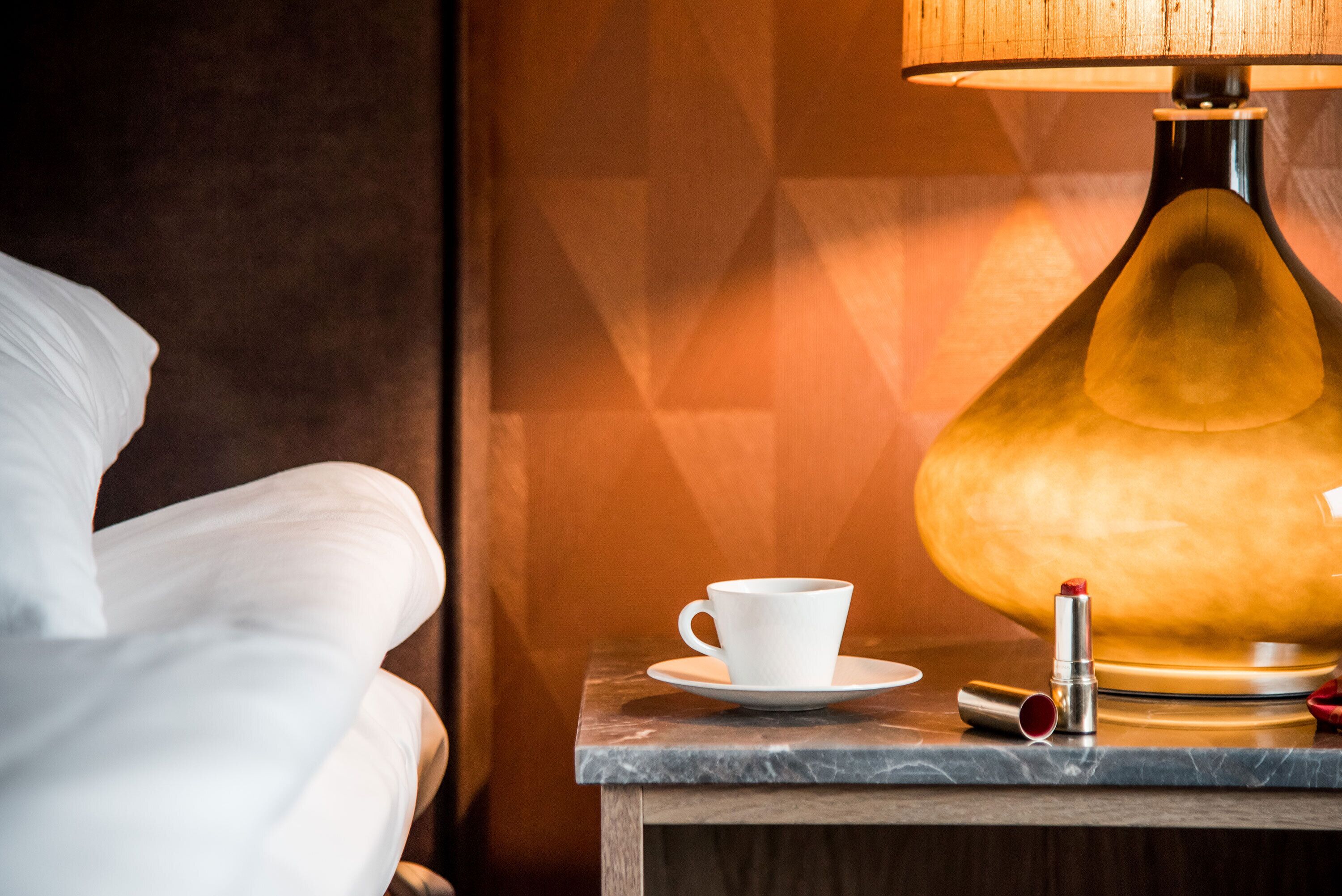 A white cup on a saucer, a lamp, and a lipstick on a wooden table.