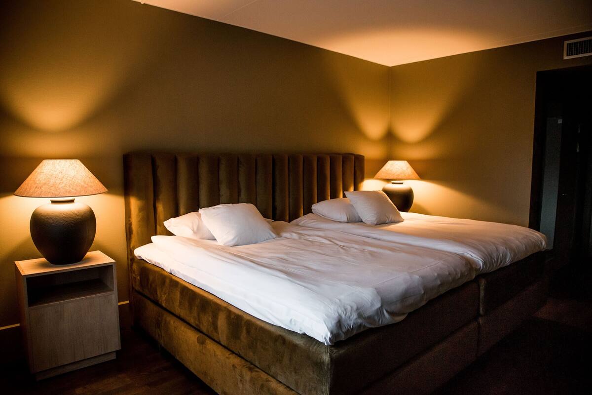 Un lit bien fait, avec du linge de lit blanc et deux oreillers, flanqué de deux tables de chevet sur lesquelles reposent des lampes, dans une pièce à l’atmosphère chaleureuse.