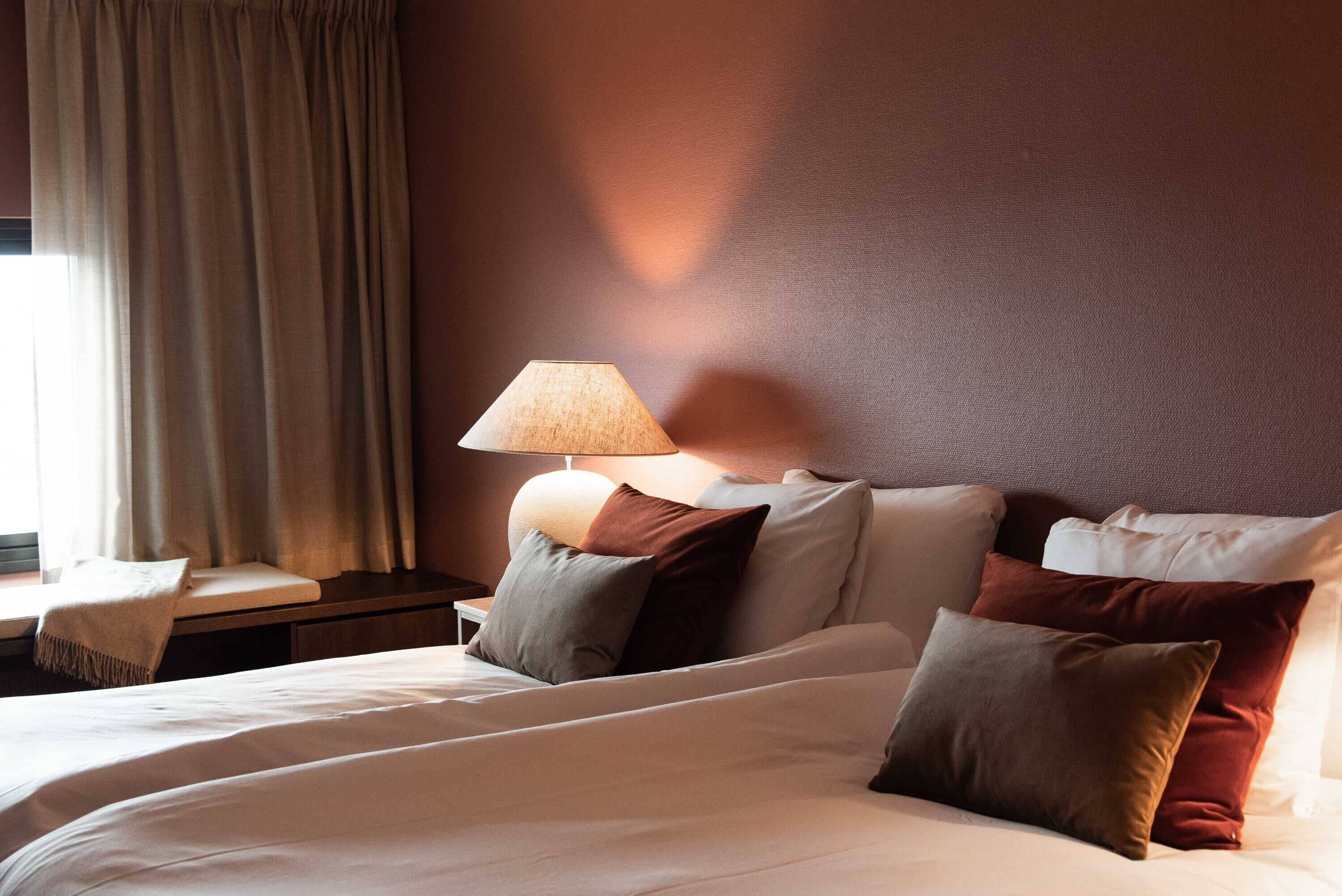Una habitación de hotel con cama, almohadas, mesita de noche y lámpara.