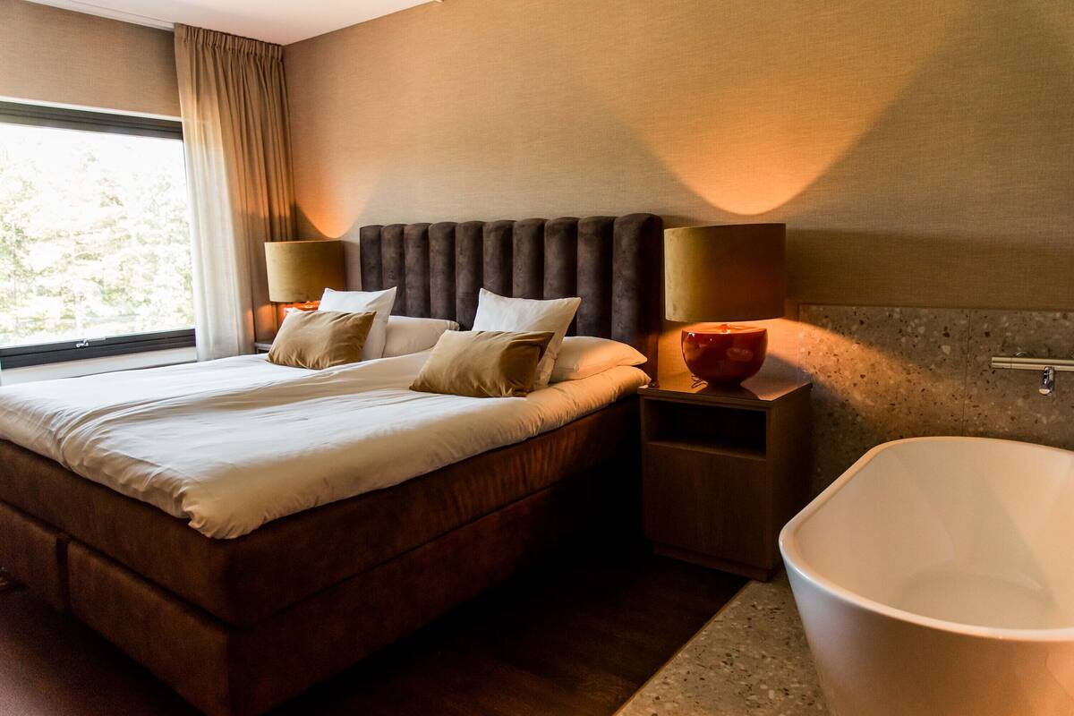 Une chambre d’hôtel moderne avec un grand lit, deux lampes de chevet, une baignoire et une fenêtre avec des rideaux.