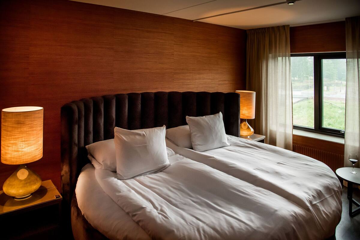 Une chambre d’hôtel avec un grand lit, deux lampes de chevet, une fenêtre avec des rideaux et une vue sur la verdure.