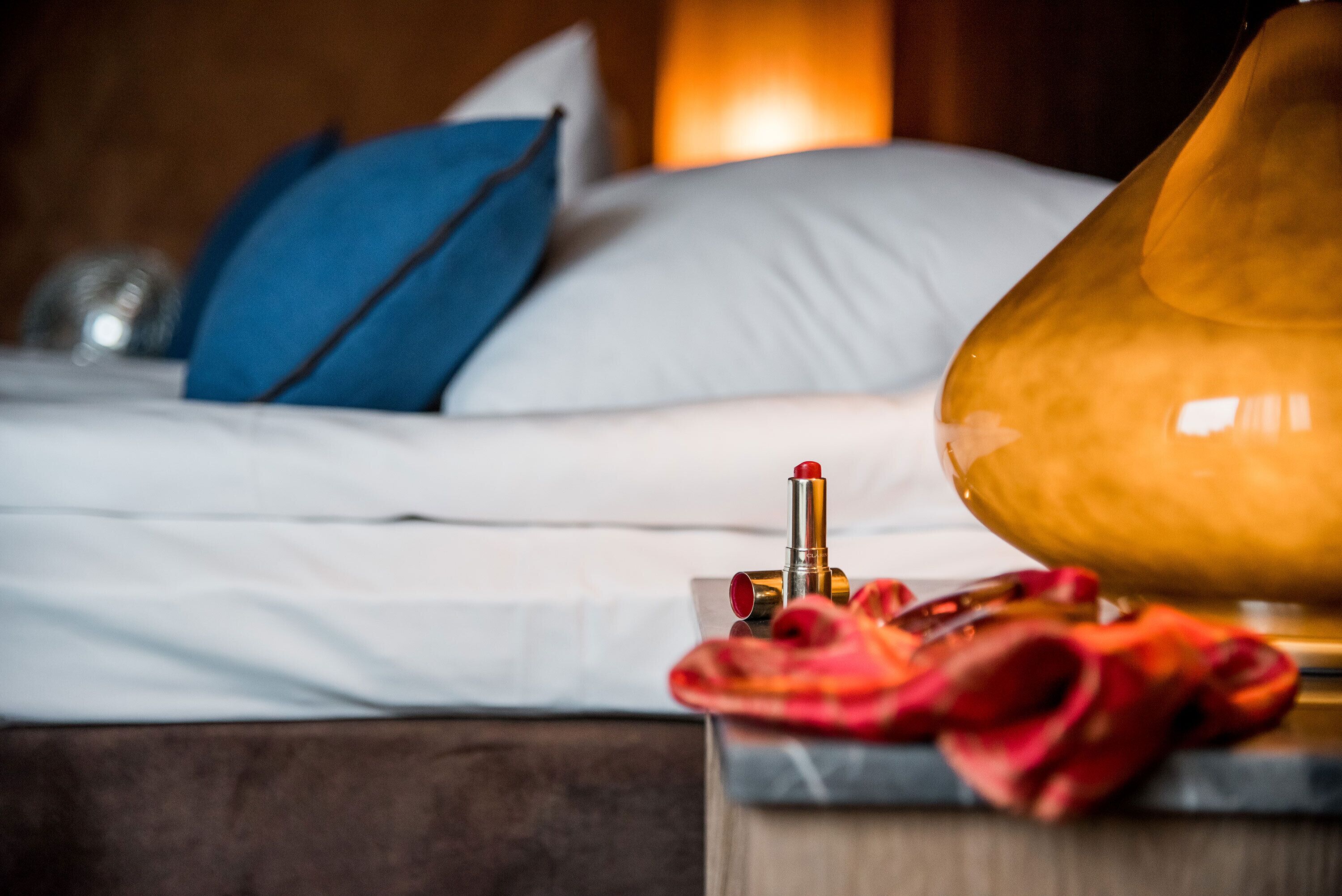 A neatly made bed with white linens and blue pillows, a bedside lamp, and a red cloth on a wooden nightstand.