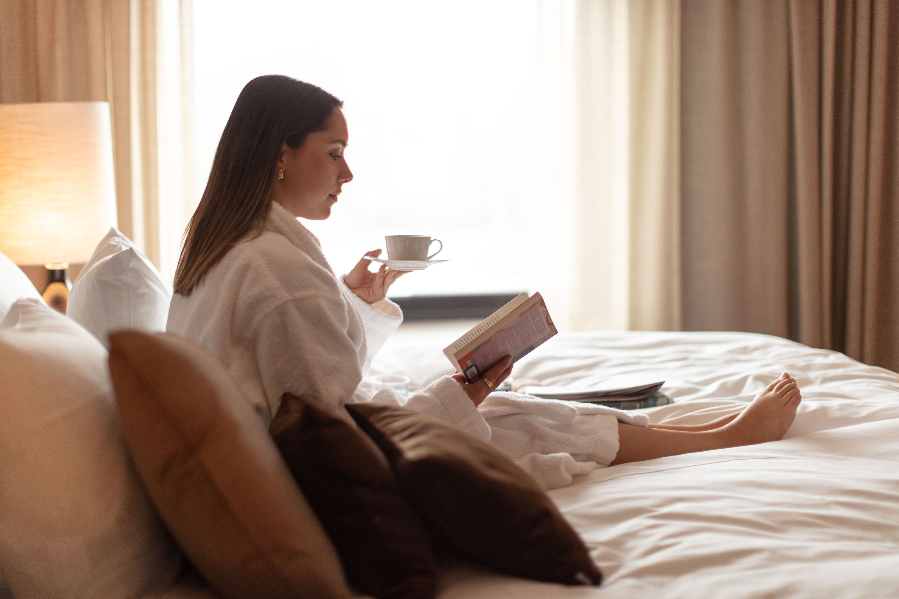 A person in a white bathrobe sitting on a bed with pillows, holding a book and a cup, in a room with a lamp and curtains.