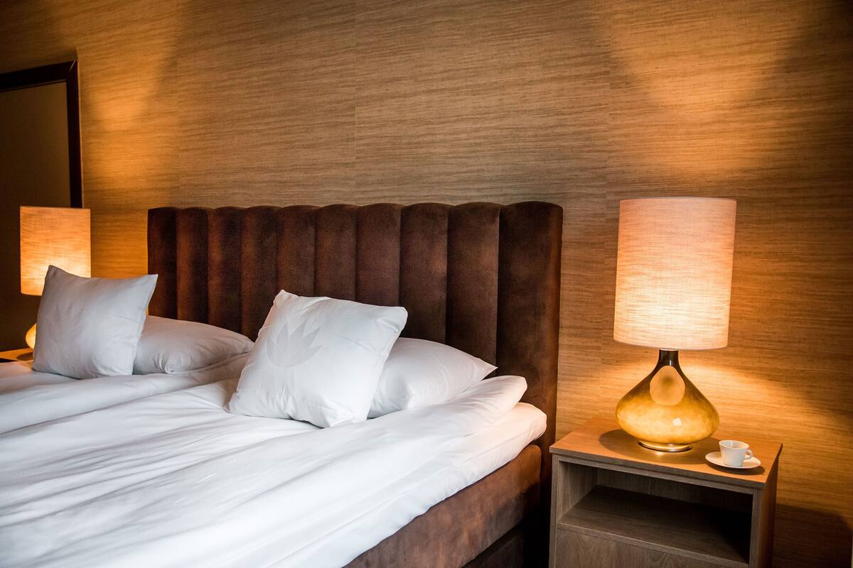 Un lit bien fait, avec du linge de lit blanc et des oreillers, une tête de lit en bois et une table de chevet sur laquelle se trouvent une lampe et une tasse.