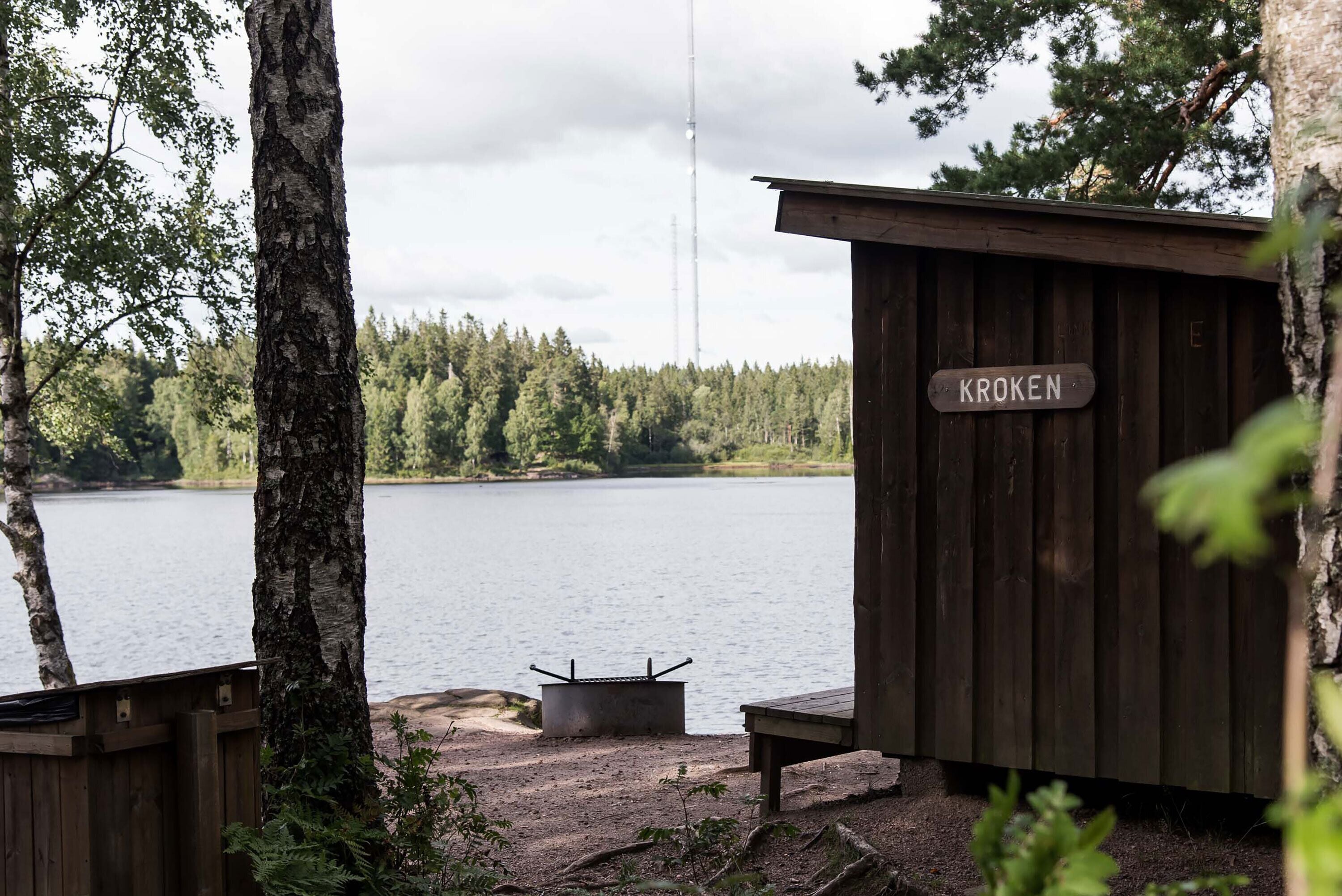 A wooden cabin with a 'KROKEN' sign, a fire pit, and a view of a lake surrounded by trees.