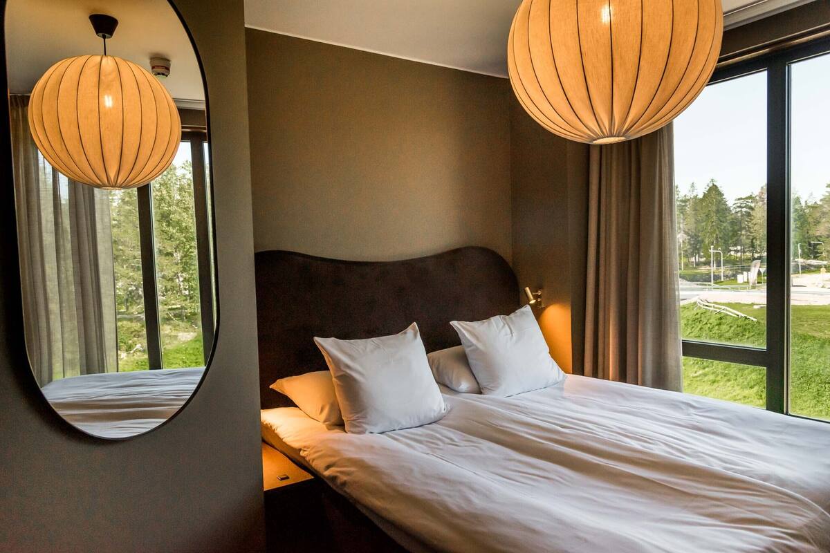 Une chambre à coucher avec un grand lit, deux oreillers blancs, une tête de lit en bois et une vue sur un parc par la fenêtre.