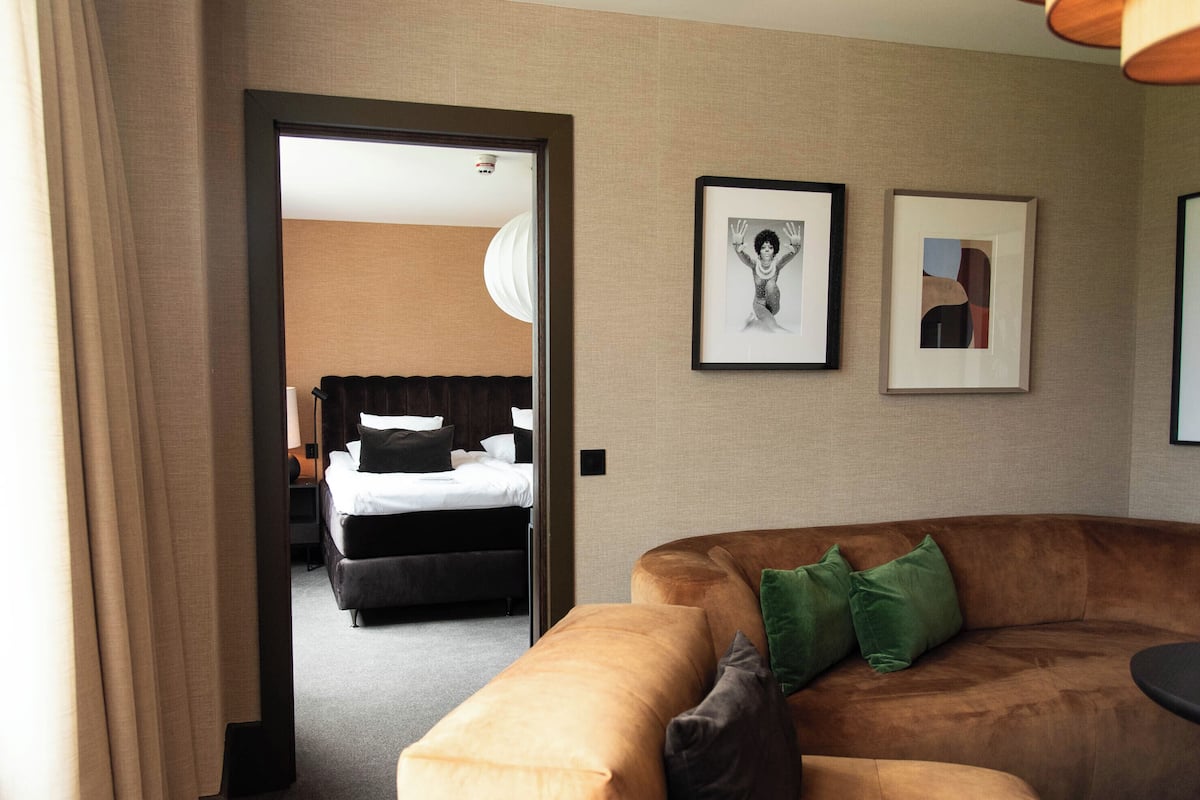 Une chambre d’hôtel avec un canapé marron, un lit avec une literie blanche et deux tableaux encadrés au mur.