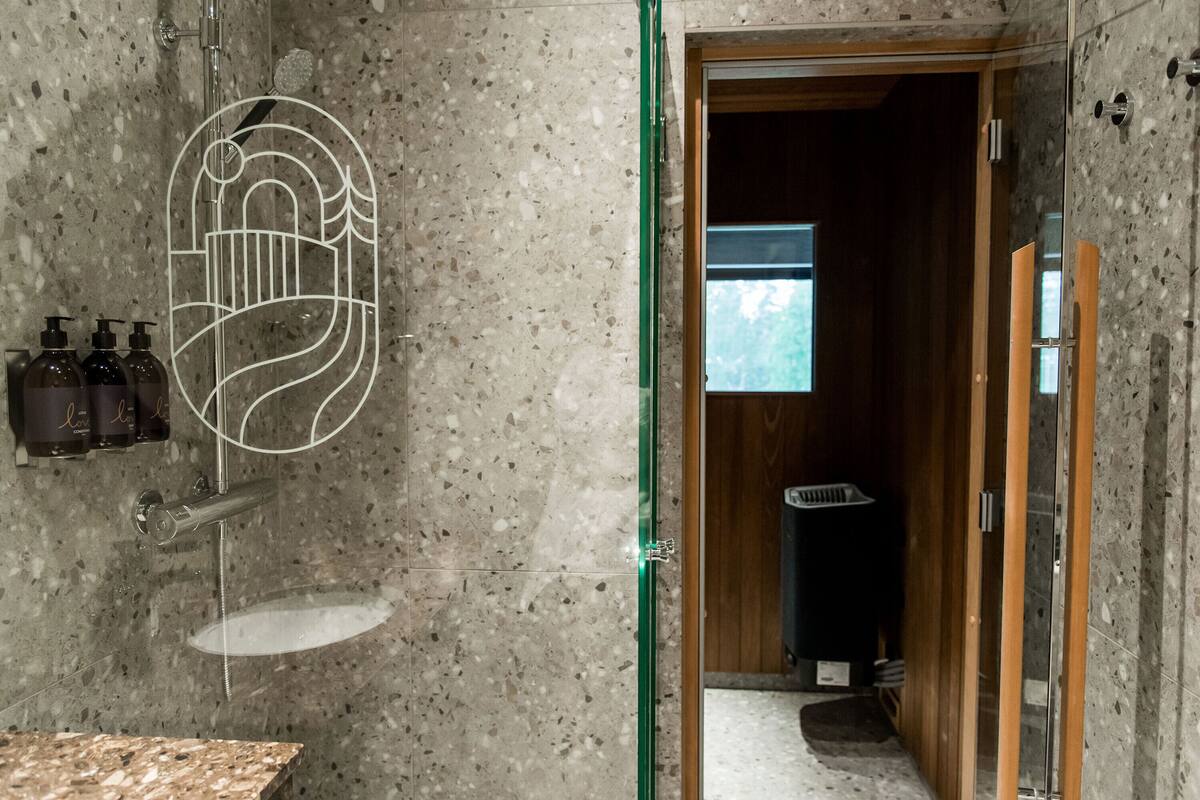 Une salle de bain dotée d’un comptoir en granit, d’une douche avec une porte en verre et d’une porte en bois donnant sur une autre pièce.