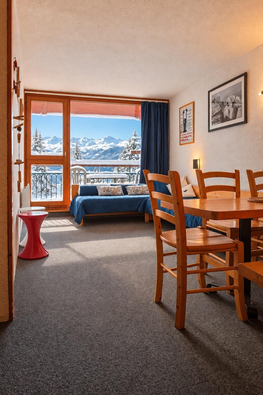 Una habitación con cama, una mesa de comedor con sillas, un taburete pequeño rojo, vistas a montañas nevadas a través de un ventanal grande y un cuadro en la pared.
