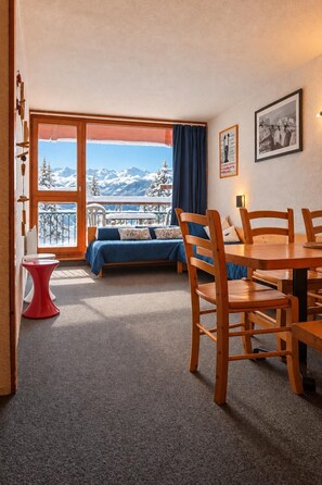 A room with a bed, a dining table with chairs, a small red stool, a view of snow-covered mountains through a large window, and a framed picture on the wall.