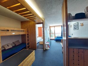 A dormitory room with bunk beds, a small seating area with stools, and a window with curtains.