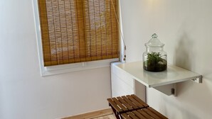 Una stanza piccola con una panca in legno, un tappeto rotondo decorato, un barattolo di vetro con piante, una lampada a parete e una finestra con tende in bambù.