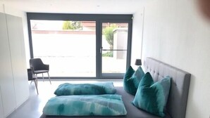 Une chambre moderne avec un canapé-lit gris, des coussins turquoise, un fauteuil et une vue sur un immeuble par le biais d’une grande fenêtre.