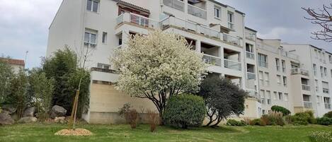 A multi-story residential building with balconies, surrounded by a garden with various shrubs and a large flowering tree.