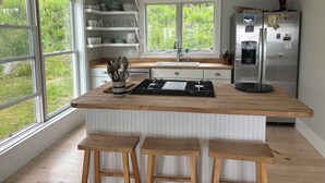 Ett modernt kök med en stor köksö, trästolar, ett kylskåp i rostfritt stål och ett fönster med utsikt över grönområden.