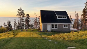 Ett envåningshus med ett mörkt tak, vita detaljer och en liten veranda. Det omges av en gräsklädd trädgård med spridda stenar och höga tallar.