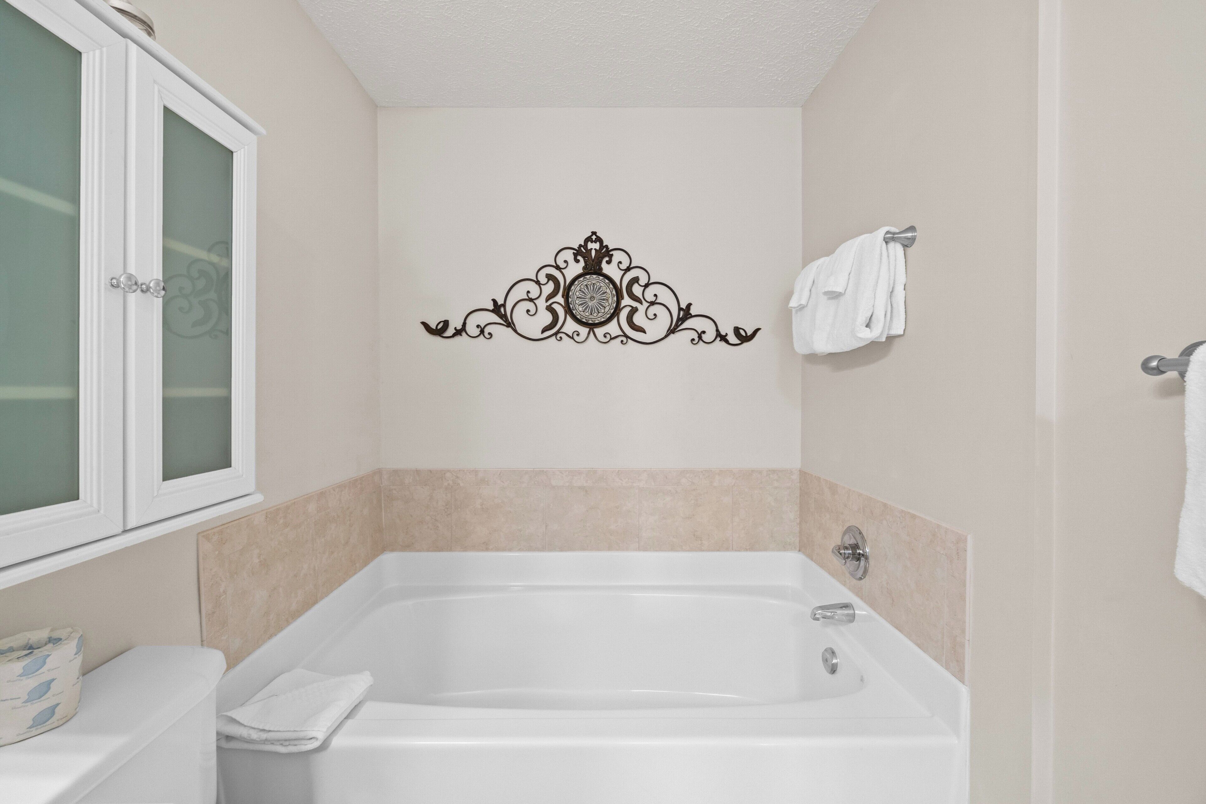 Een badkamer met een wit bad, beige tegels, een decoratief wandobject en een raam met gezandstraald glas.
