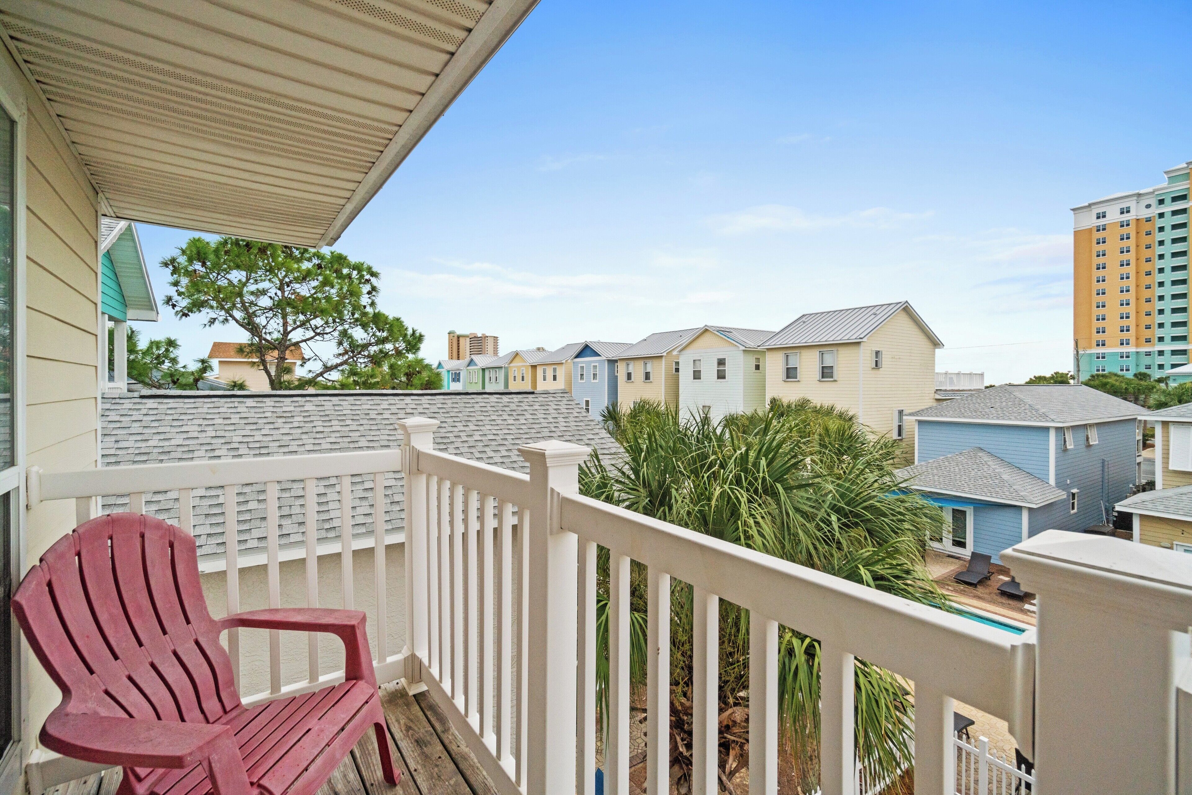 A balcony with a red Adirondack chair, a white railing, and a view of neighboring houses and a tall building in the background.