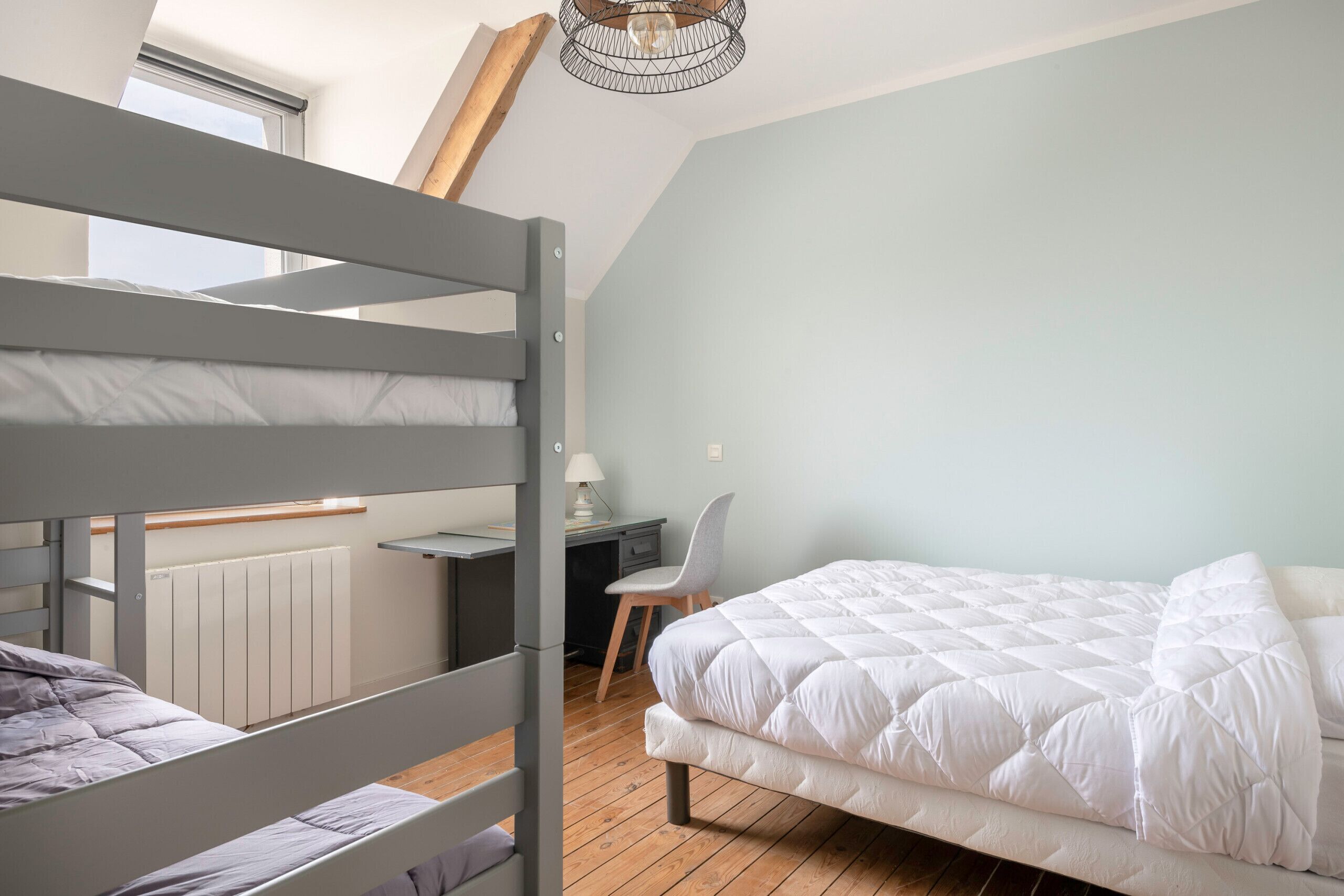 A dormitory room with bunk beds, a desk, a chair, and a large window.