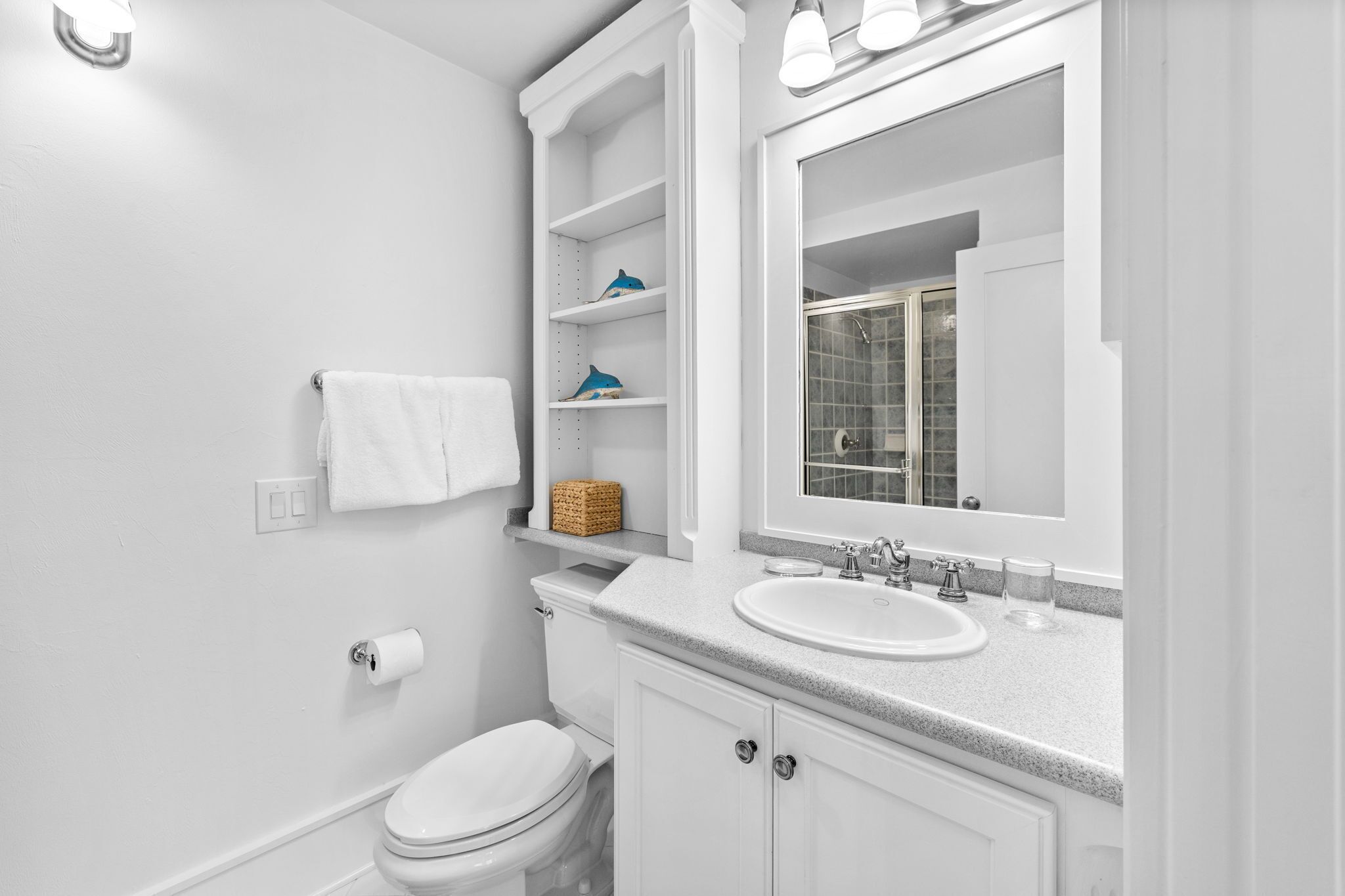 A white bathroom with a toilet, a sink, a mirror, and shelves with toiletries.