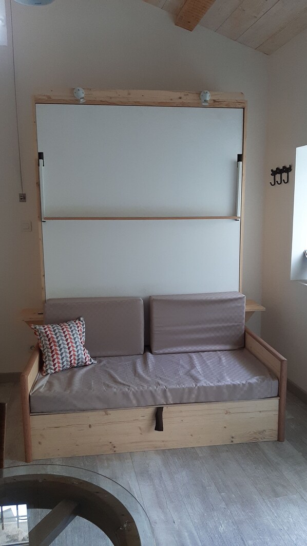 Un canapé en bois avec deux coussins et un oreiller à motifs, placé contre un mur où se trouve un interrupteur et un petit objet décoratif.