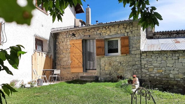 A stone building with a wooden door and window shutters, a small garden with a metal chair, and a metal fence. - Cosy studio in a farmhouse  (UBRAYE)