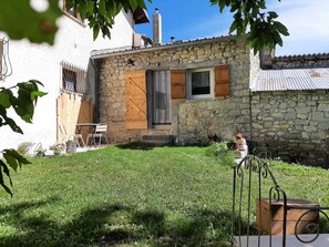 Un bâtiment en pierre avec une porte et des volets en bois, un petit jardin avec une chaise en métal et une clôture en métal.