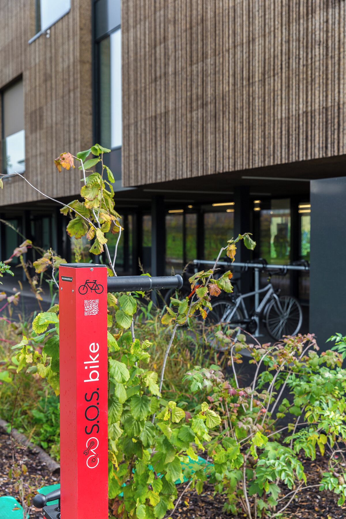 A red bike parking sign with a QR code and a bicycle symbol, located in front of a modern building with large windows and a bicycle parked inside.