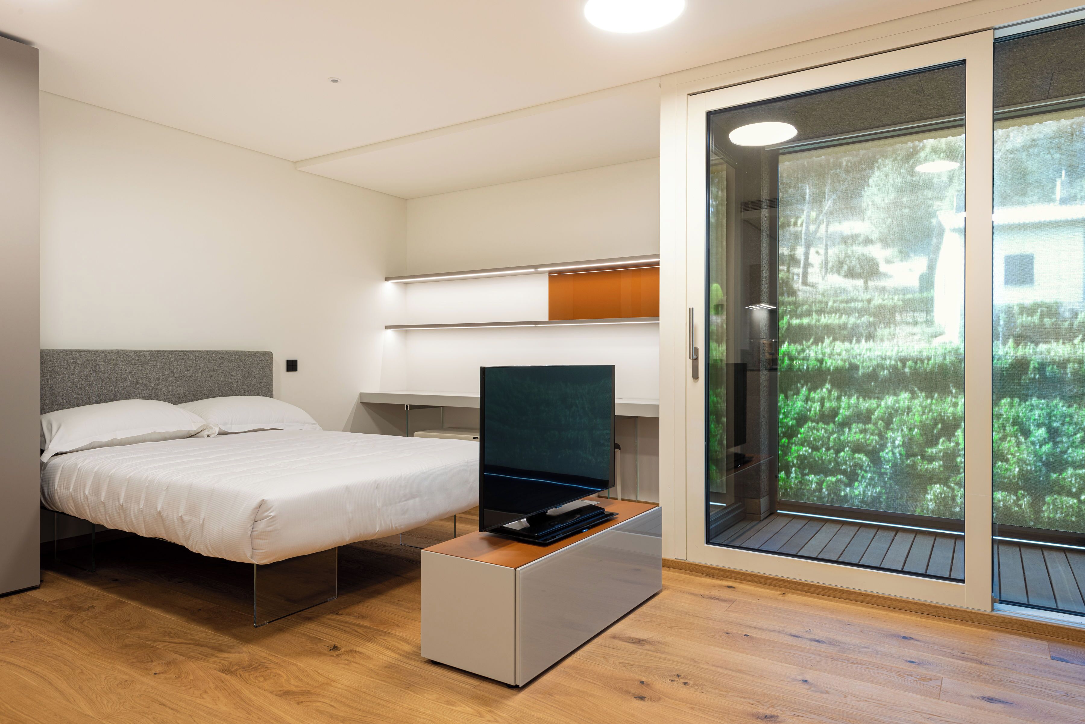 A modern bedroom with a bed, a flat-screen TV on a wooden stand, a desk with a chair, and a sliding glass door leading to a garden.