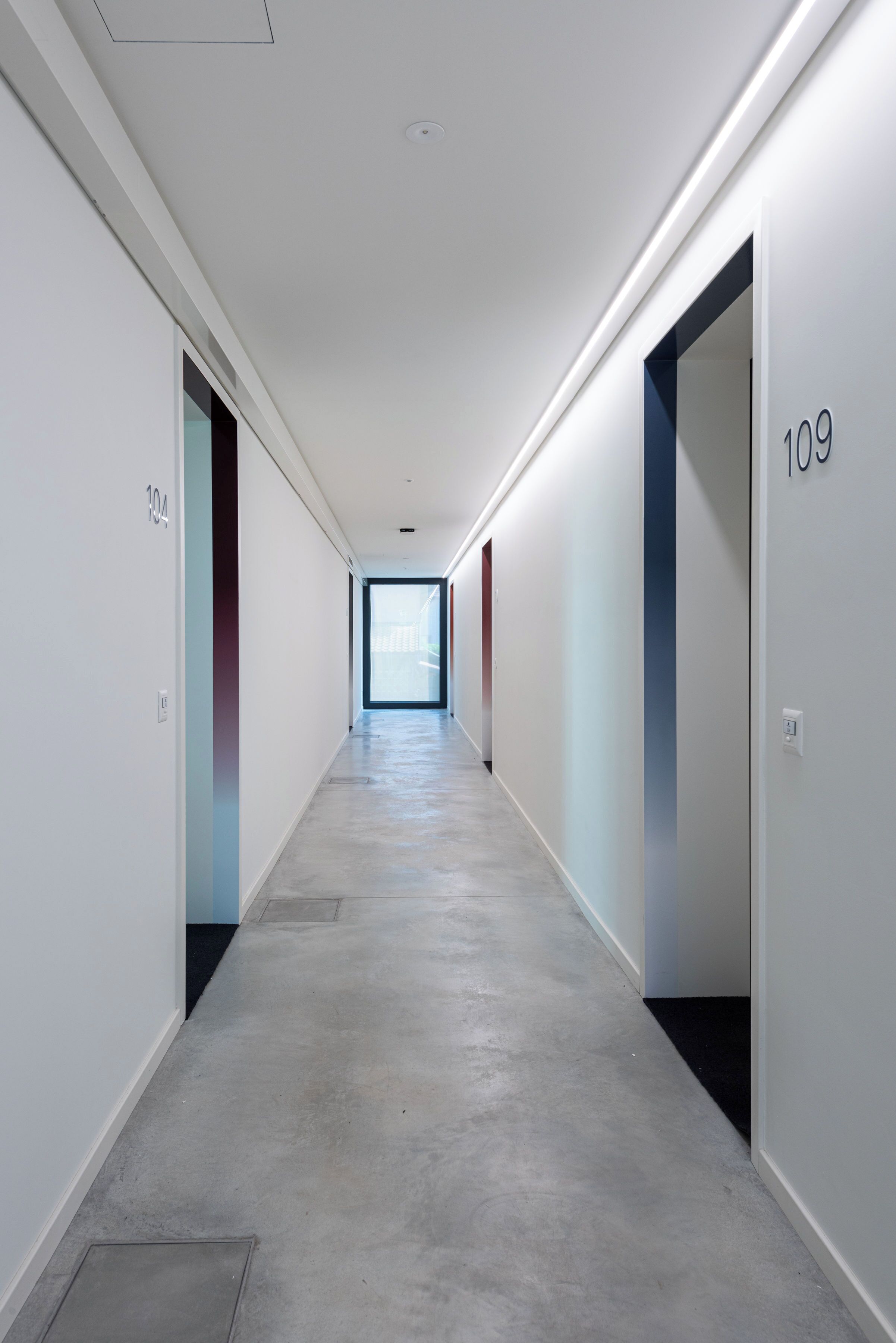 Un pasillo largo y estrecho con paredes blancas y suelo gris, que conduce a una habitación luminosa al final.