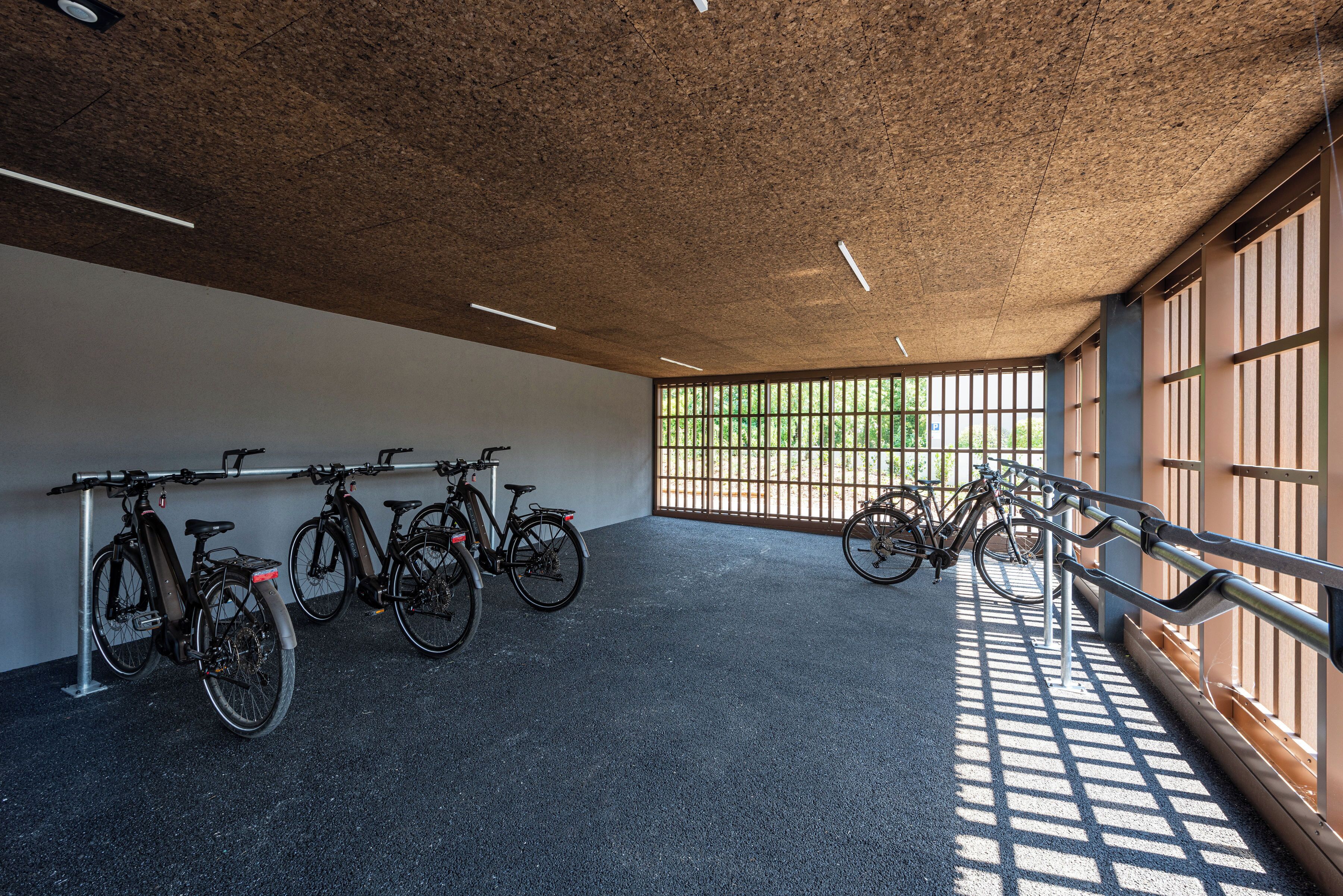 Et overdækket cykelparkeringsområde med cykler, beton gulv og store vinduer der lukker naturligt lys ind.