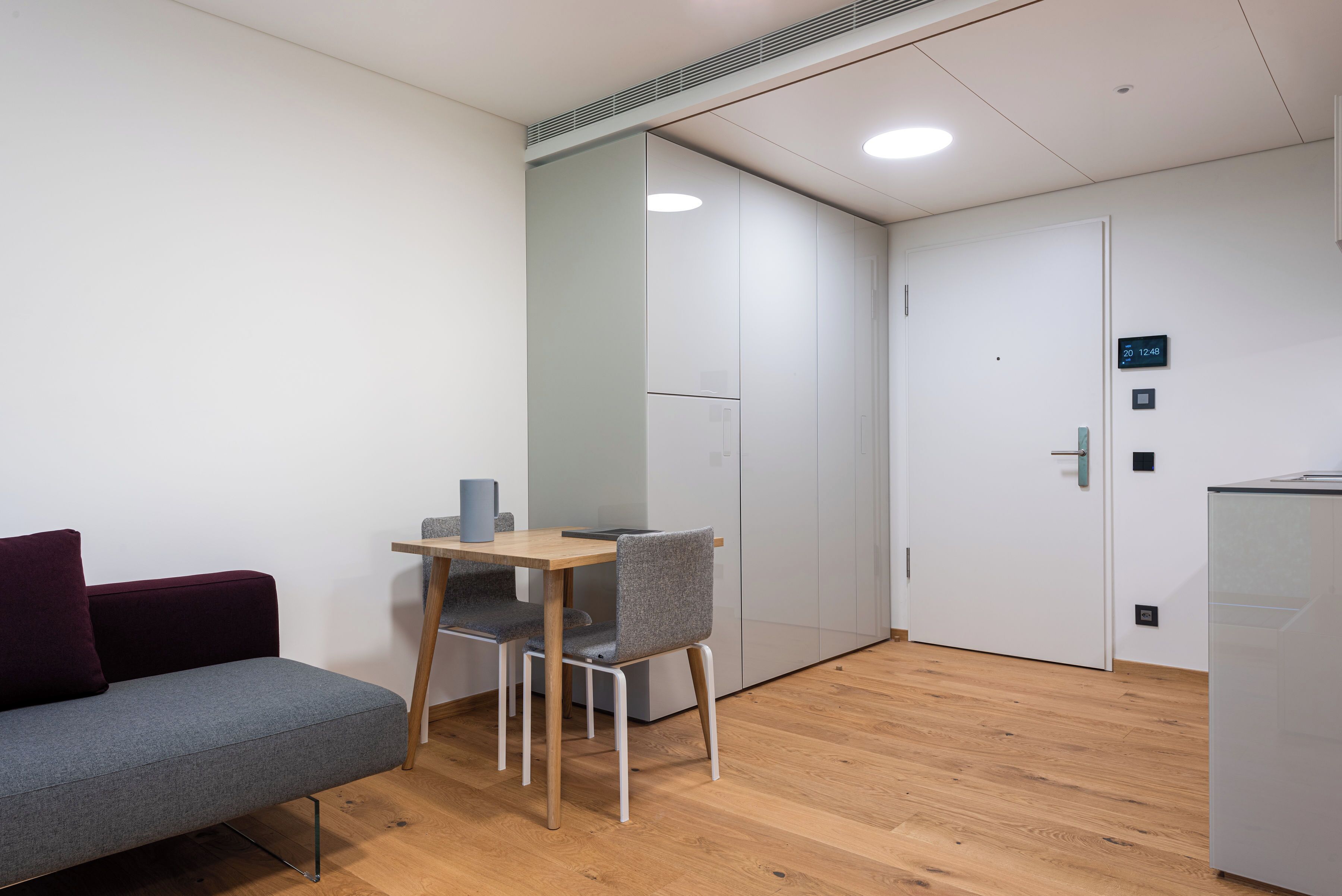 A modern room with a grey sofa, a small wooden table, and a grey chair. There is a white door with a digital clock and a small cabinet.