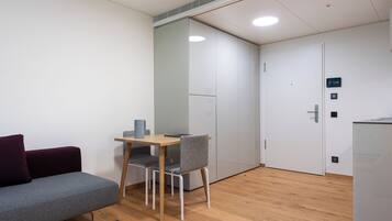 A modern room with a grey sofa, a small wooden table, and a grey chair. There is a white door with a digital clock and a small cabinet.