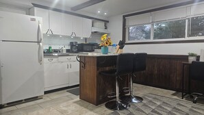 Une cuisine avec des meubles blancs, un îlot central en bois, un tabouret de bar et une fenêtre avec des stores.