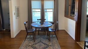 Une salle à manger avec une table ronde, quatre chaises, un lustre et une vue par une fenêtre équipée de stores.