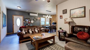 A cozy living room with a brown leather sofa, a wooden coffee table, and a ceiling fan. There is a dining area with a table and chairs, and a kitchen in the background.