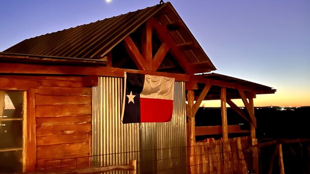 A wooden cabin with a corrugated metal door and a Texas flag hanging on the front.