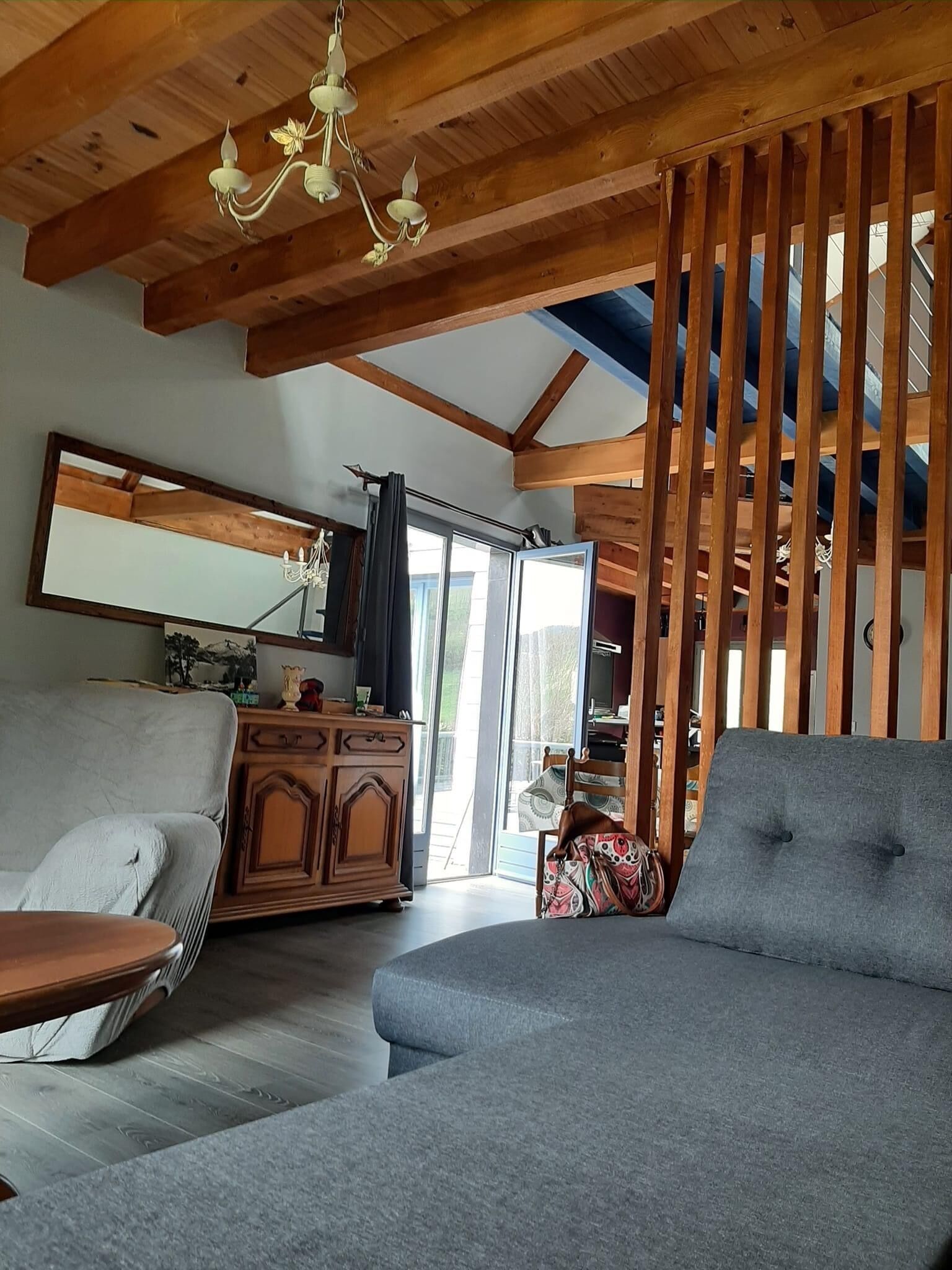 Een ruime woonkamer met een hoge plafond, houten balken, een kroonluchter, een grijze bank, een houten kast en een grote spiegel.