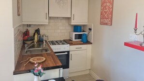 A compact kitchen with white cabinets, a gas stove, a microwave, and a sink. There is a decorative chandelier, a red candle holder, and a small potted plant on the counter. - Lisheens Lodge (Kealkill)