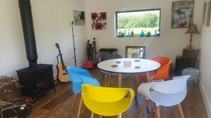 A cozy room with a round table, colorful chairs, a guitar, a wood-burning stove, and a window with a view of bottles. - Lisheens Lodge (Kealkill)