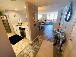 A modern kitchen with white cabinets, a stainless steel sink, and a dishwasher. There is a dining area with a table and chairs, and a living room with a sofa and a coffee table.
