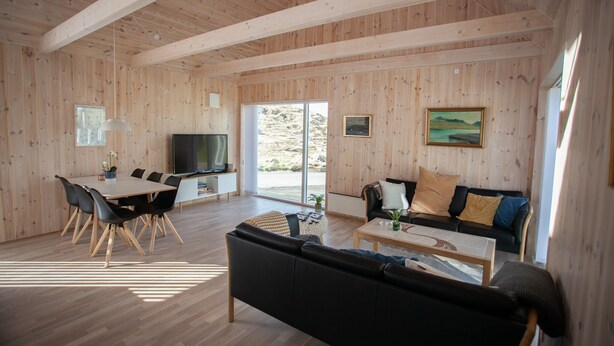 A spacious living area with a wooden ceiling, a dining table with chairs, a black sofa, a coffee table, and a television. - Vacation Home - Scenic Nature - Sandoy (Sandur)