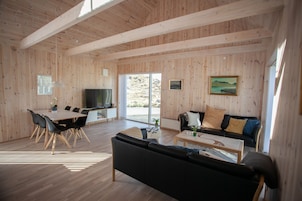 A spacious living area with a wooden ceiling, a dining table with chairs, a black sofa, a coffee table, and a television.