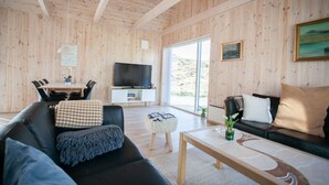 A modern living room with a wooden interior, a flat-screen TV, a dining area with a table and chairs, and a sofa with cushions. - Vacation Home - Scenic Nature - Sandoy (Sandur)