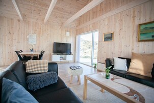 A modern living room with a wooden interior, a flat-screen TV, a dining area with a table and chairs, and a sofa with cushions.