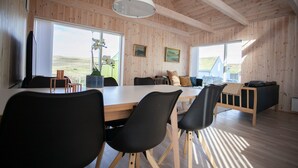 A spacious room with a large wooden table and chairs, a hanging light fixture, and large windows offering a view of the outdoors. - Vacation Home - Scenic Nature - Sandoy (Sandur)