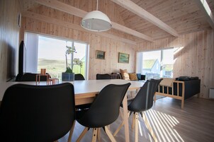 A spacious room with a large wooden table and chairs, a hanging light fixture, and large windows offering a view of the outdoors.