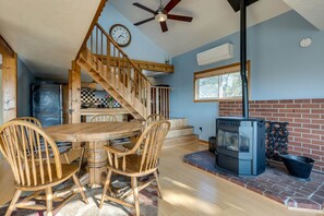 Un comedor con mesa y sillas de madera, una cocina con refrigerador y estufa, una escalera y un ventanal con vistas a árboles.