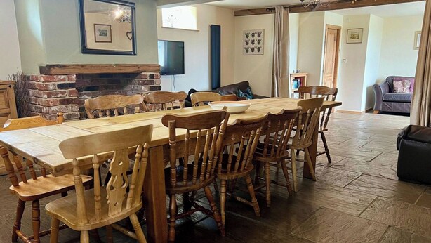 Un espace repas comprenant une longue table en bois et des chaises, une cheminée en briques et un téléviseur fixé au mur.